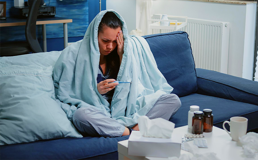 Mulher sentada no sofá, enrolada em um cobertor e com um termômetro na mão. Tem remédios e papel toalha na mesinha em frente a ela.